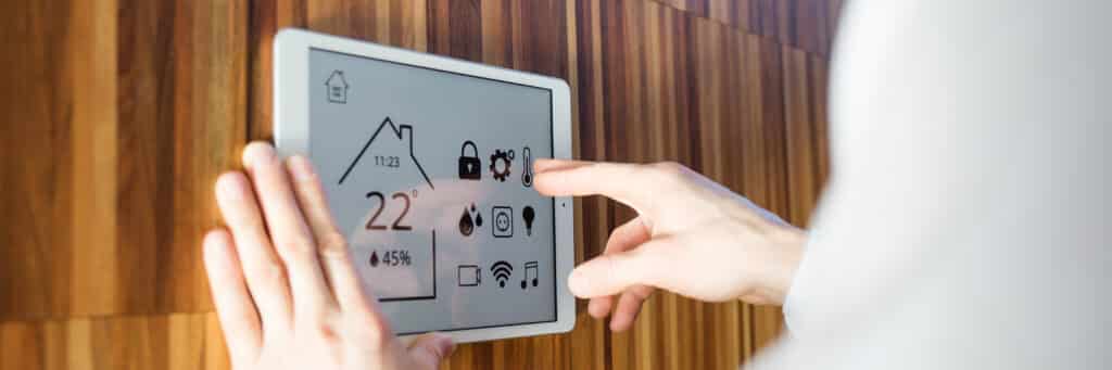 Main contrôlant la domotique sur une tablette murale Smart Home. L'écran affiche 22°C, 45% humidité, sécurité, lumières, WiFi. Mur en bois.