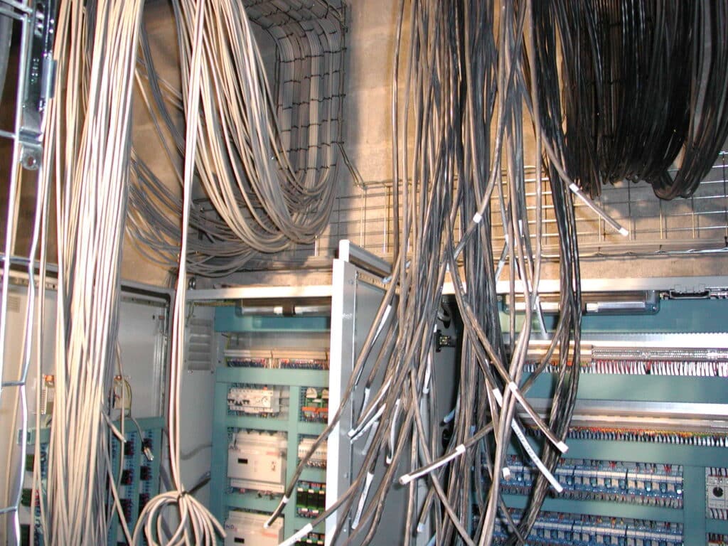 Câblage électrique industriel : organisation et contrôle Câblage électrique industriel : des chemins de câbles muraux supportent de nombreux câbles gris vers des armoires de commande ouvertes, révélant disjoncteurs et borniers.