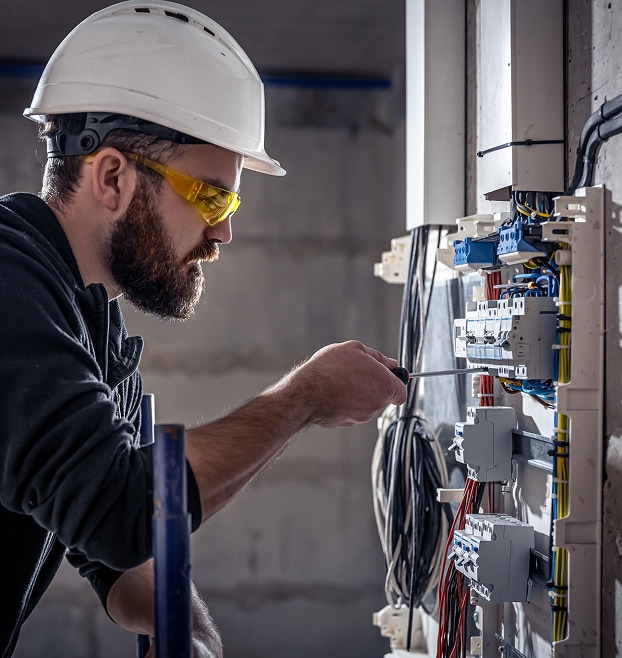 Électricien barbu en casque blanc et lunettes de sécurité jaunes, vissant des fils sur un panneau électrique avec disjoncteurs.