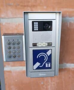 Interphone vidéo Golmar argenté sur mur de briques. Clavier numérique, caméra, haut-parleur. Écran: "LUTINS | PRINCE". Symbole d'aide auditive.