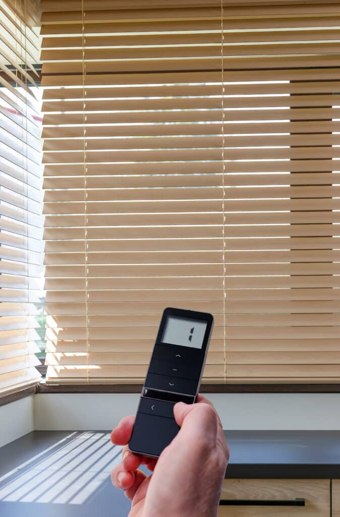 Main contrôlant des stores en bois avec une télécommande affichant '1'. Le soleil filtre à travers les lamelles, créant des ombres sur le comptoir.