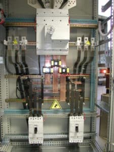 Intérieur d'armoire électrique : disjoncteurs, barres de cuivre, transformateurs de courant, câblage et signalisations danger électricité. ON/OFF visible.