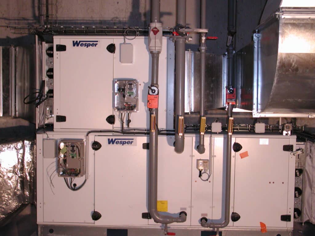 Wesper CVC : Unités de Traitement d'Air Industrielles Unité de traitement d'air modulaire Wesper en salle mécanique. Panneaux blancs, tuyaux gris, vannes et conduits électriques. Connectée à des gaines métalliques.